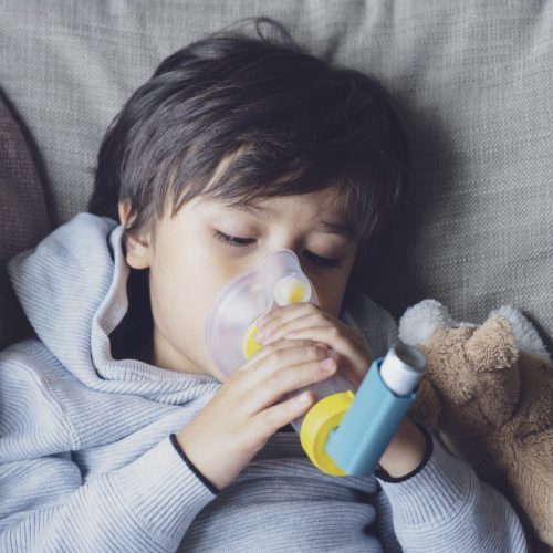 Poor boy tired from chest coughing holding inhaler mask, Child closing his eyes while using the volumtic for breathing treatment,Tried Kid having asthma allergy using the asthma inhaler