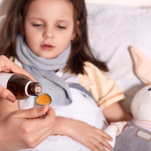 Horizontal photo of unknown hand pouring liquid into spoon from little bottle with syrup, person caring about child lying in bed with flu and sore throat, being upset. Children and health concept.